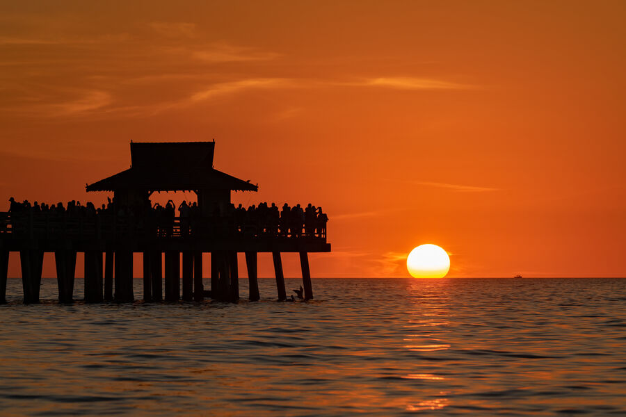 Fine art print of Naples Pier at nautical twilight, dock lights beginning to glow as the sky transitions from orange to deep indigo above the Gulf